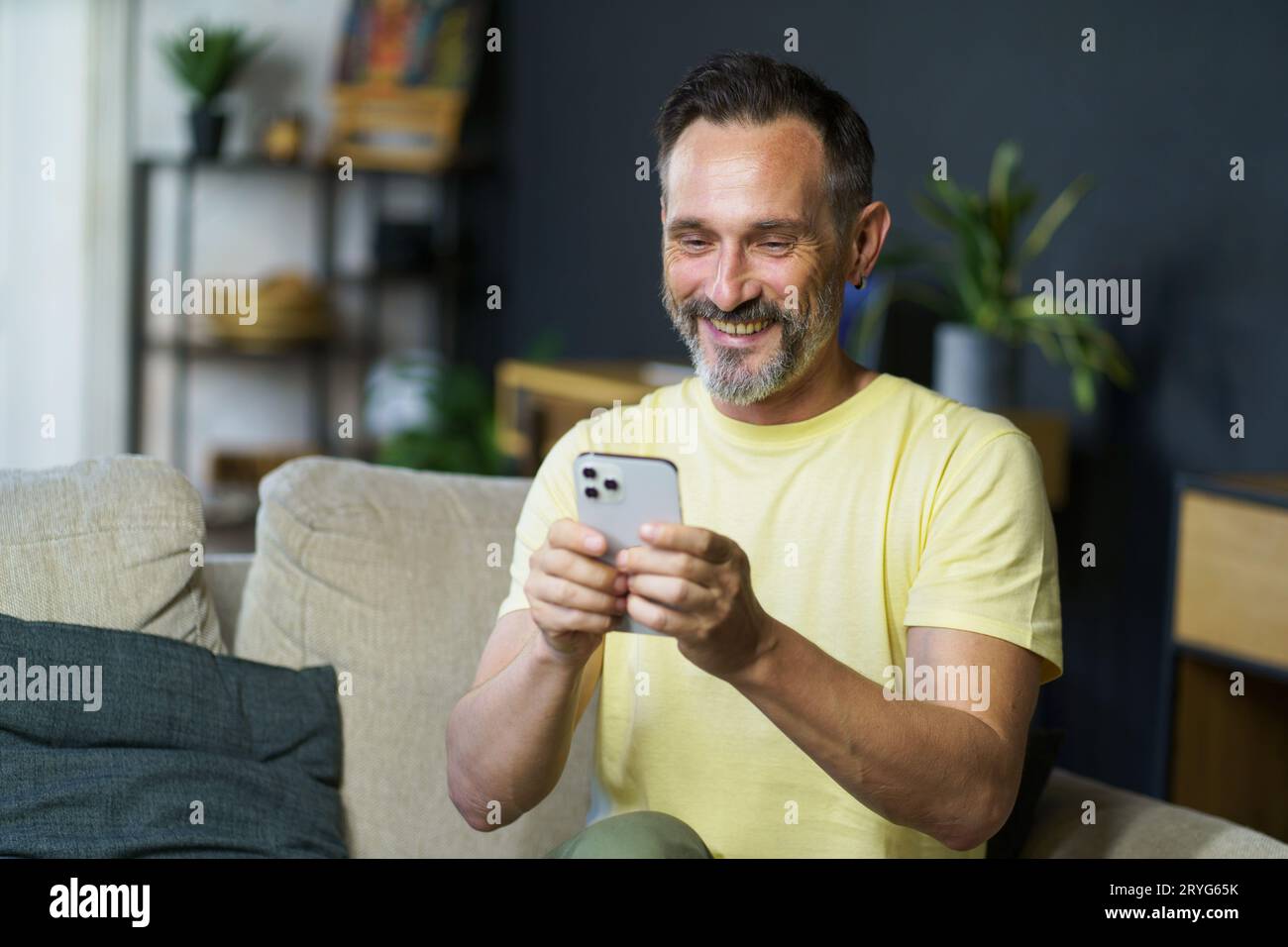 L'affascinante uomo di mezza età usa lo smartphone per inviare e-mail, controllando i social media seduti sul divano all'interno di casa. Uomo maturo sen Foto Stock