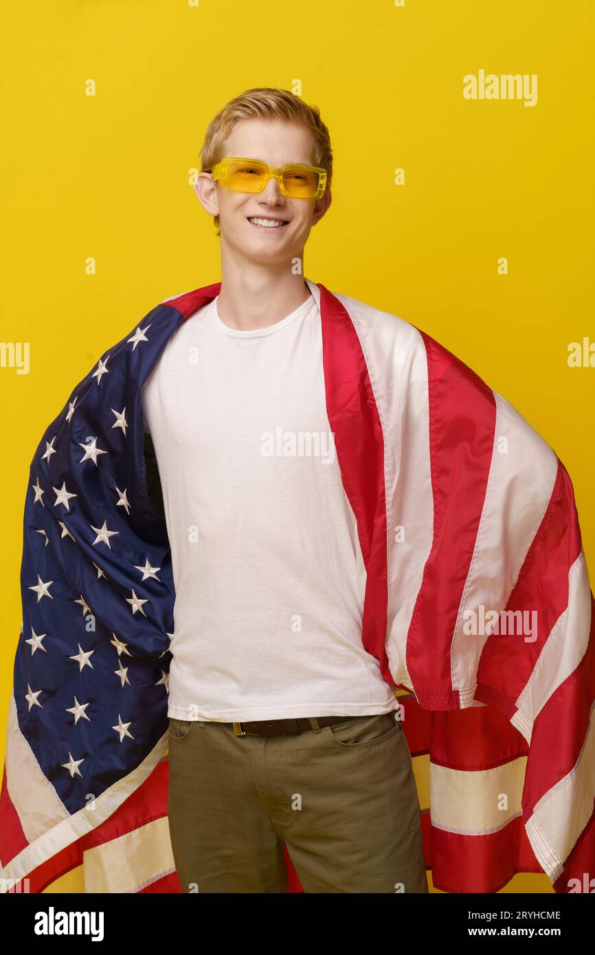 Super American Hero Concept. Youth Smiles in una maglietta bianca e la bandiera americana è isolata su uno sfondo giallo. Foto Stock