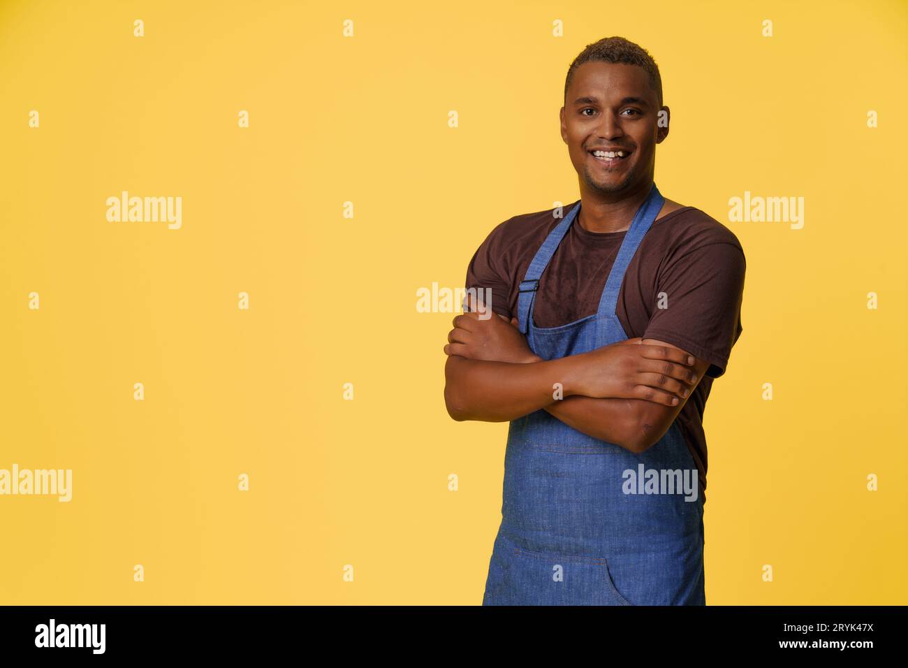 Allegro cuoco afro-americano che indossa un grembiule blu sorride di fronte a sfondo giallo con spazio per la copia, suggerendo il concetto di pr Foto Stock