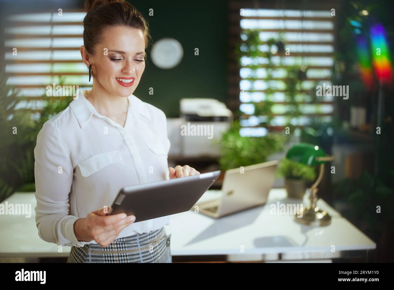 Un'attività immobiliare sostenibile. Donna d'affari felice ed elegante di mezza età in un moderno ufficio verde in camicetta bianca che utilizza un tablet PC. Foto Stock