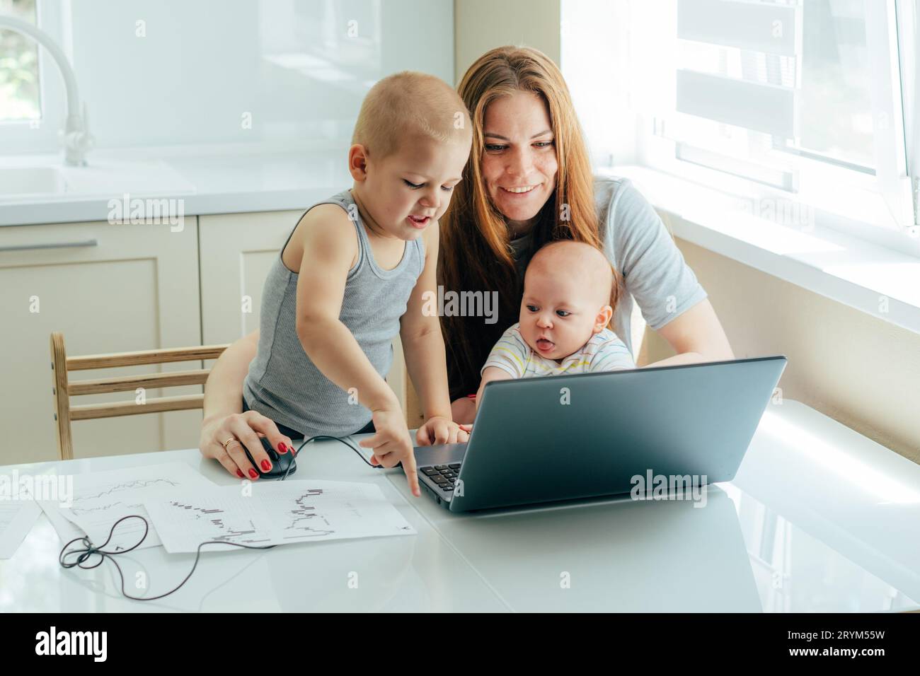 Giovane attraente madre con due bambini studia online su un computer portatile in cucina a casa. Foto Stock