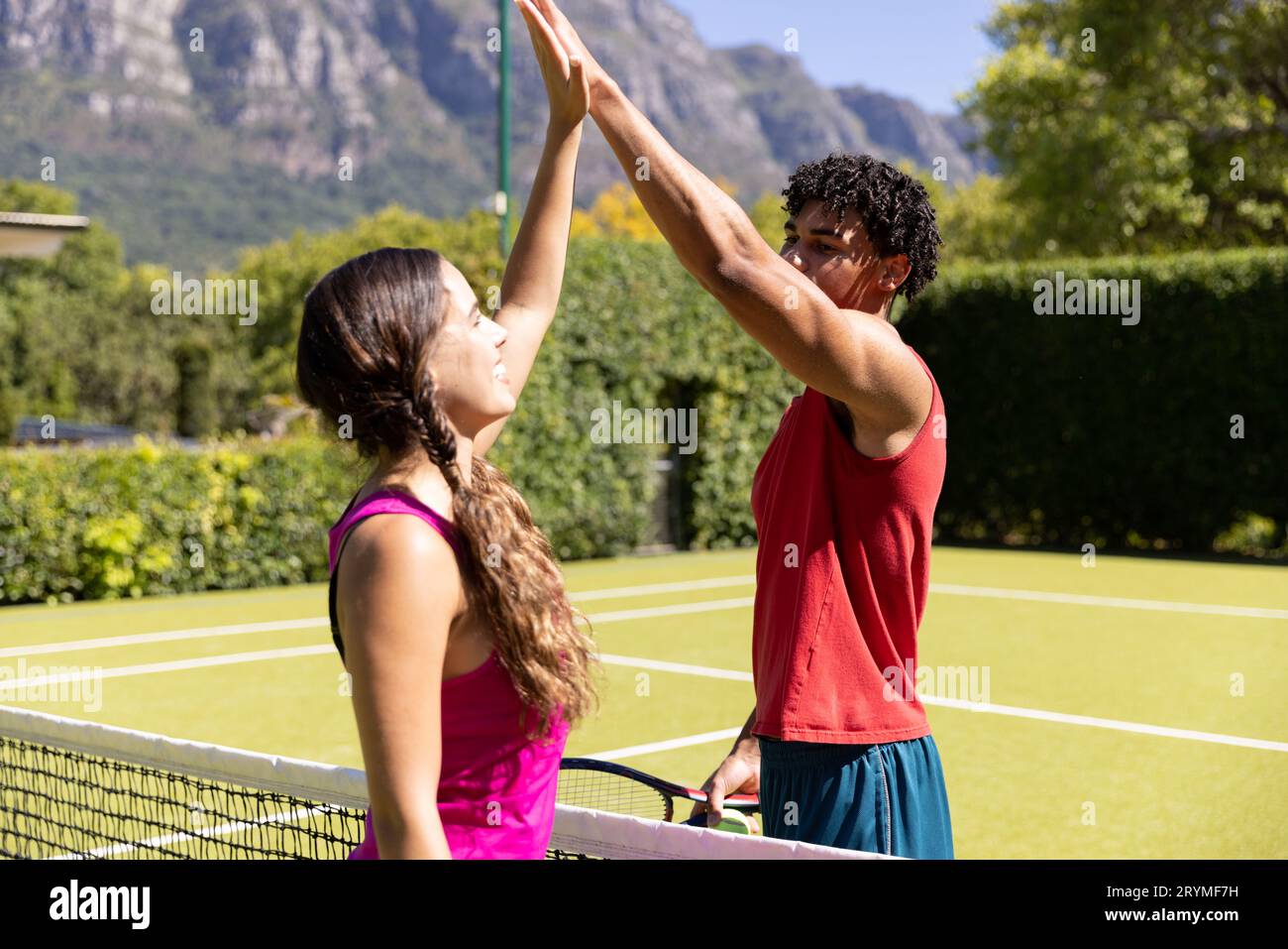 Felice coppia diversa reggendo racchette, alto fiving su rete sul soleggiato campo da tennis all'aperto Foto Stock