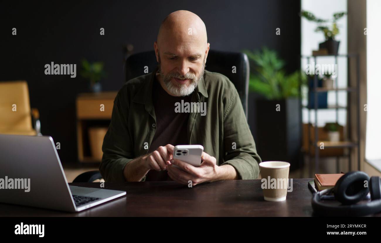 Uomo anziano felice e carismatico che si è impegnato in messaggi di testo al telefono mentre si prendeva una pausa dal lavoro con un notebook. Uomo anziano, pieno Foto Stock