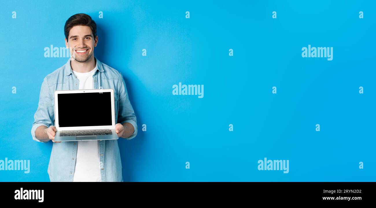 Bello giovane uomo introdurre il prodotto sullo schermo del laptop, mostrando il computer e sorridendo, in piedi su sfondo blu Foto Stock