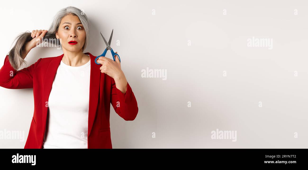 Donna anziana asiatica che tiene forbici e guarda nervosa alla macchina fotografica, pensa a tagliare i capelli, cambiare acconciatura, stare in piedi Foto Stock
