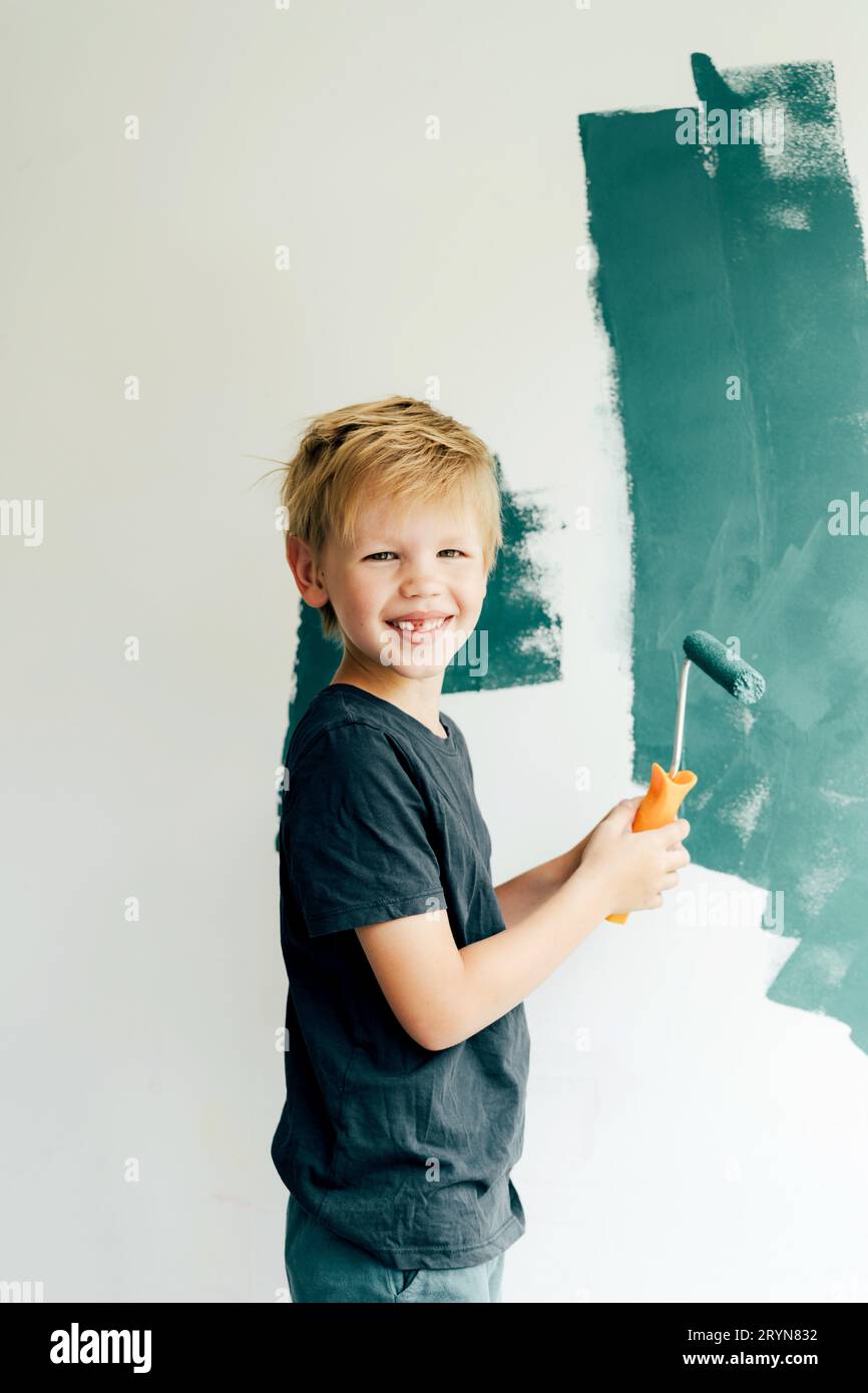 Il biondo caucasico dipinge un muro bianco con un rullo in verde. Il bambino guarda la macchina fotografica e sorride un sorriso senza denti. A sc Foto Stock