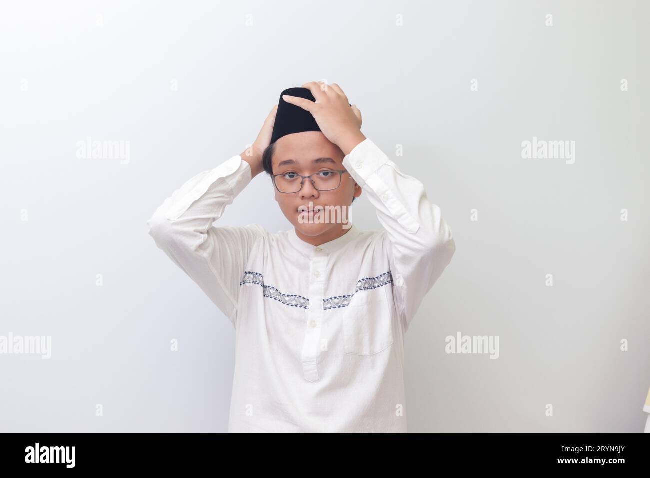 Ritratto di un giovane musulmano asiatico che cerca di regolare il suo songkok o Skullcap nero. Immagine isolata su sfondo bianco Foto Stock