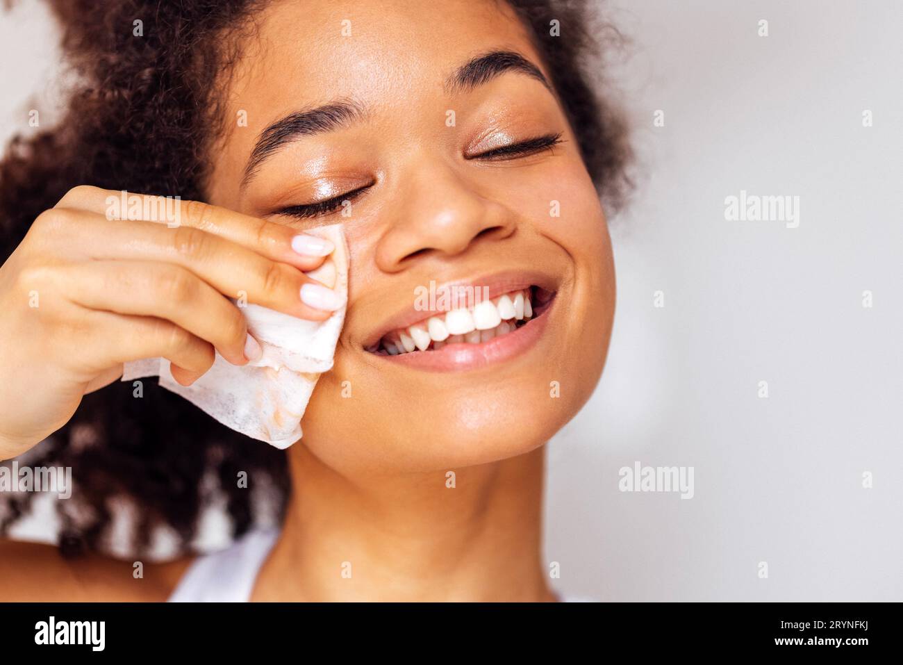 Primo piano del volto della giovane felice e attraente africana che si lava il trucco. Affascinante ragazza di razza mista sorridente si pulisce la pelle del viso Foto Stock