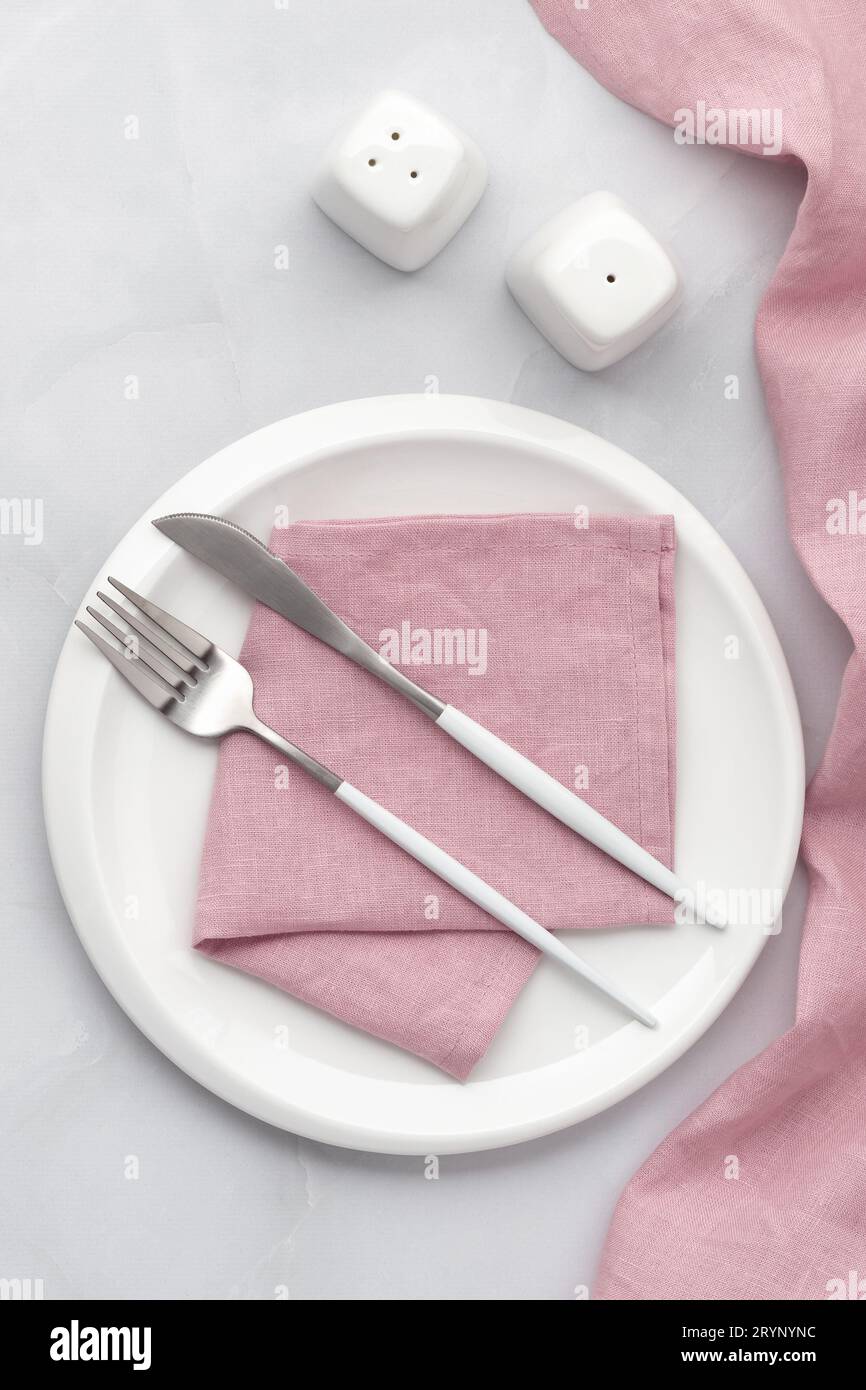Piatto bianco con posate in argento. Coltello, forchetta e tovagliolo rosa su sfondo grigio. Impostazione del piano d'esame, piano d'esame, vista dall'alto. Spazio per servire, piatto in ceramica pulito. Foto Stock