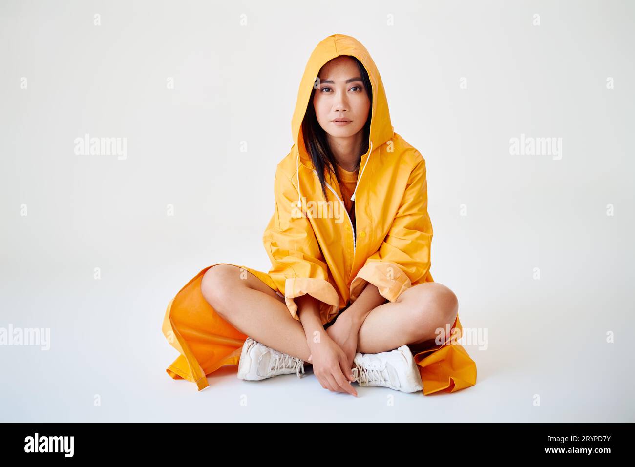 Sorridente ragazza asiatica con bretelle vestite con un impermeabile giallo brillante in posa con cappuccio sulla testa su sfondo bianco dello studio seduta sul pavimento con una copia di spumante Foto Stock