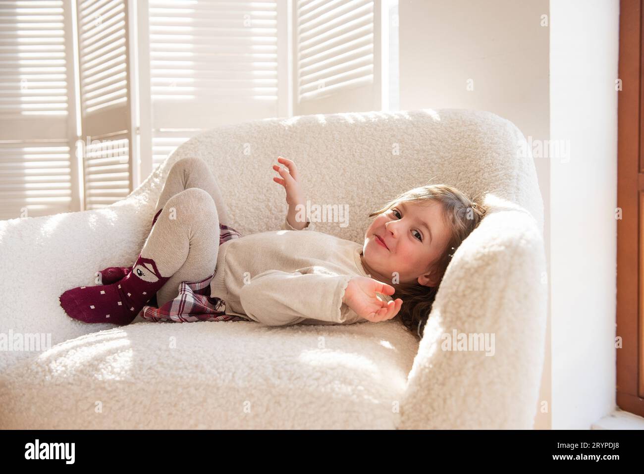Simpatica bambina giace su una morbida sedia bianca, ingannando, facendo facce vicino all'albero di Natale. Bambino in un'atmosfera accogliente a casa. Interi festivi Foto Stock