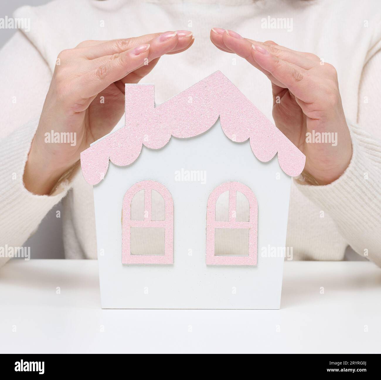 Le mani femminili si piegano l'una all'altra su una casa in miniatura in legno su sfondo bianco. Concetto di assicurazione immobiliare, envi Foto Stock