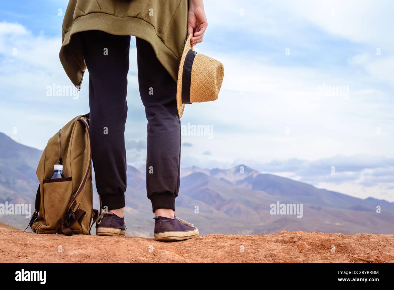 Una ragazza turistica si trova sulla collina. Gambe del viaggiatore, cappello e zaino con bottiglia d'acqua da vicino Foto Stock