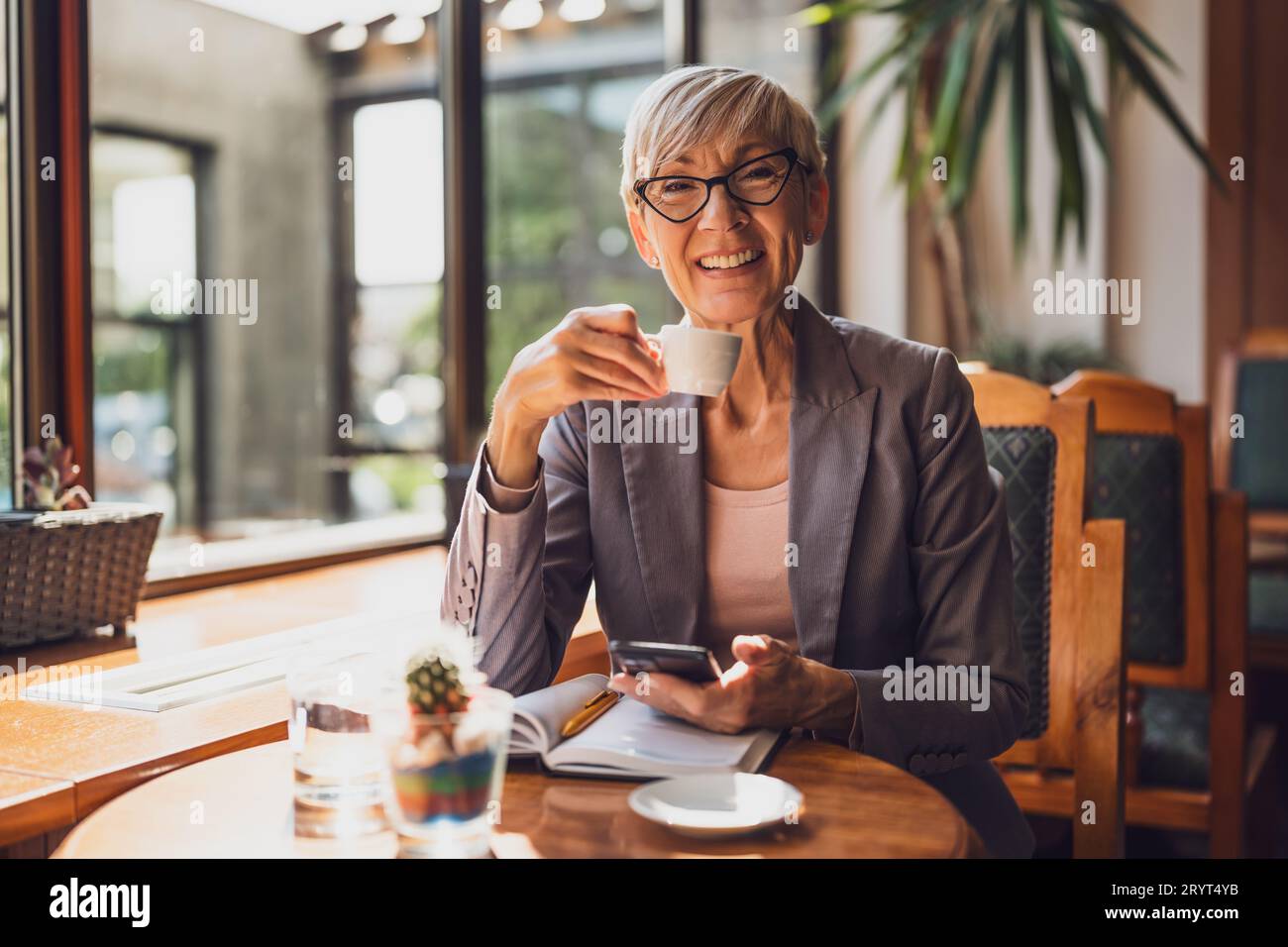 Una donna matura è seduta in una caffetteria e si sta rilassando. Sta bevendo caffè e sta usando lo smartphone. Foto Stock