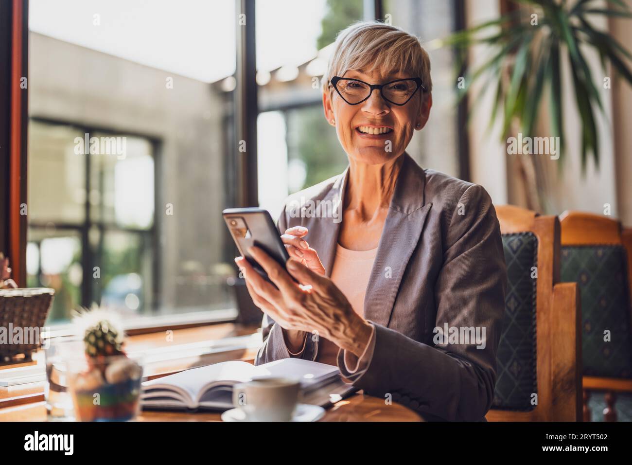 Una donna matura è seduta in una caffetteria e si sta rilassando. Sta bevendo caffè e sta usando lo smartphone. Foto Stock