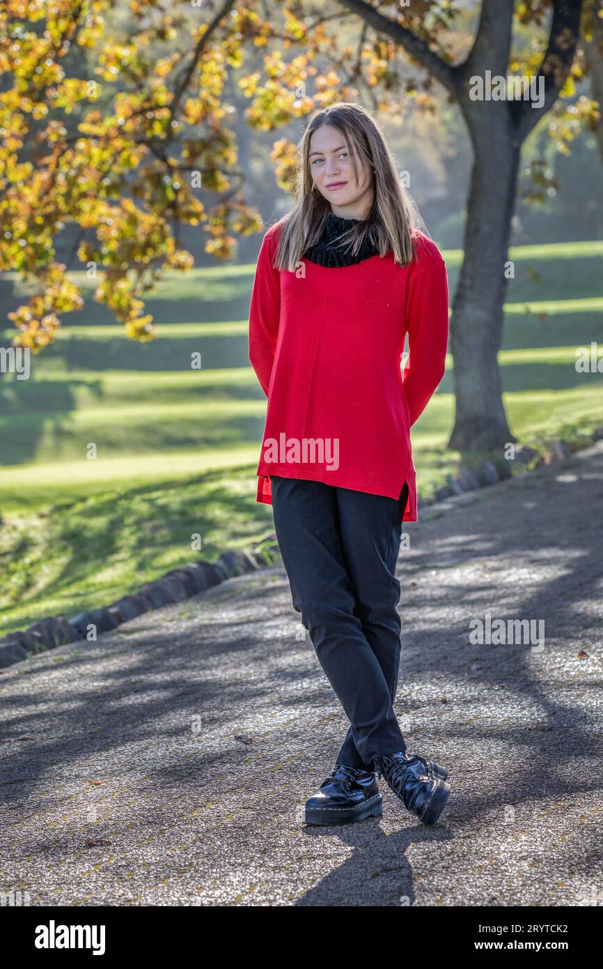 In un colpo alla testa e alle spalle, una splendida giovane donna bionda, che indossa un cappello di lana nera e un maglione rosso brillante, si irradia sotto gli alberi autunnali Foto Stock