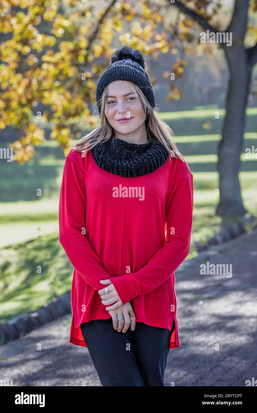 Una bella giovane donna bionda, che indossa un cappello di lana nera e un vivace maglione rosso, si erge incantatamente sotto gli alberi autunnali Foto Stock