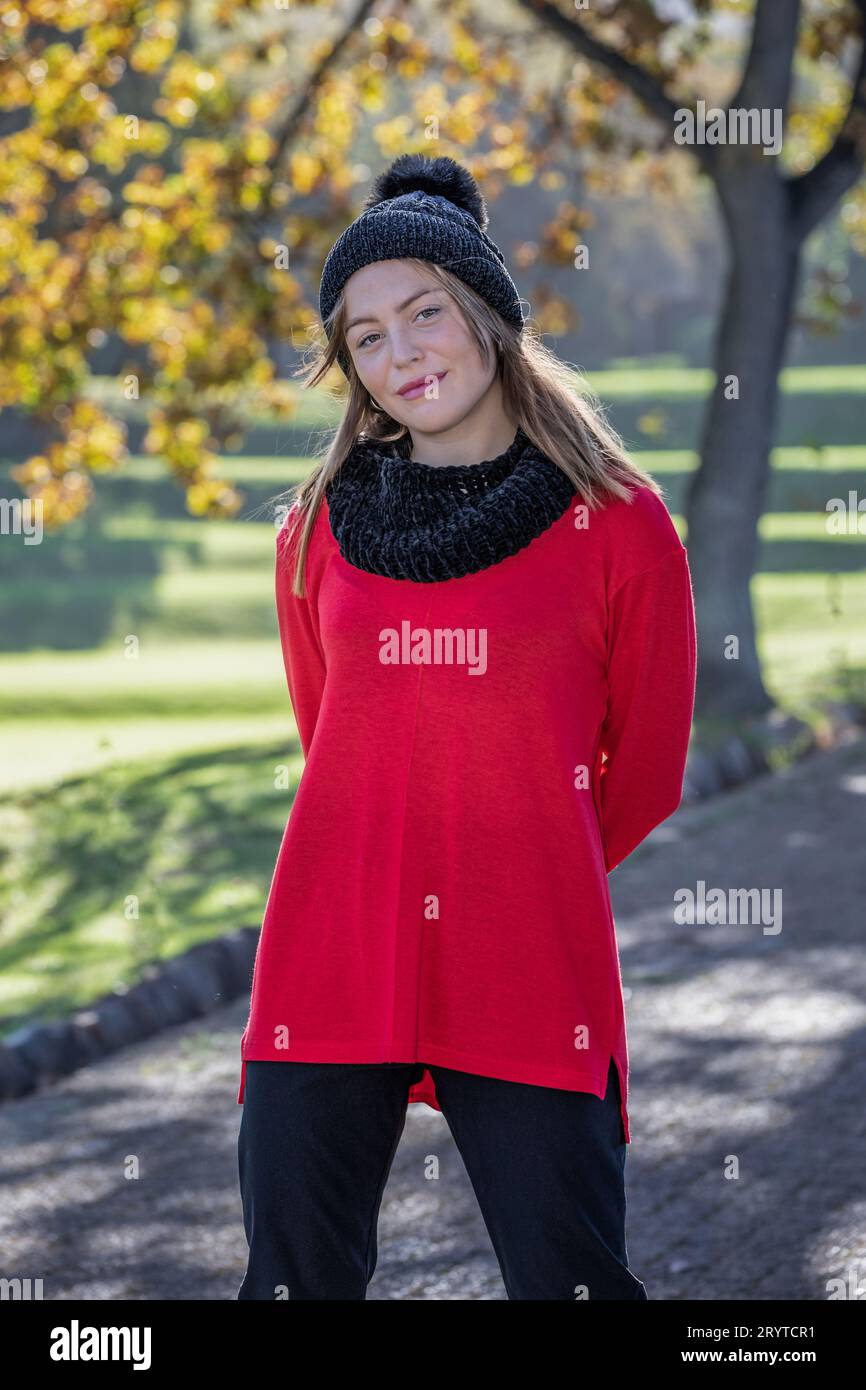Una bella giovane donna bionda, che indossa un cappello di lana nera e un vivace maglione rosso, si erge incantatamente sotto gli alberi autunnali Foto Stock