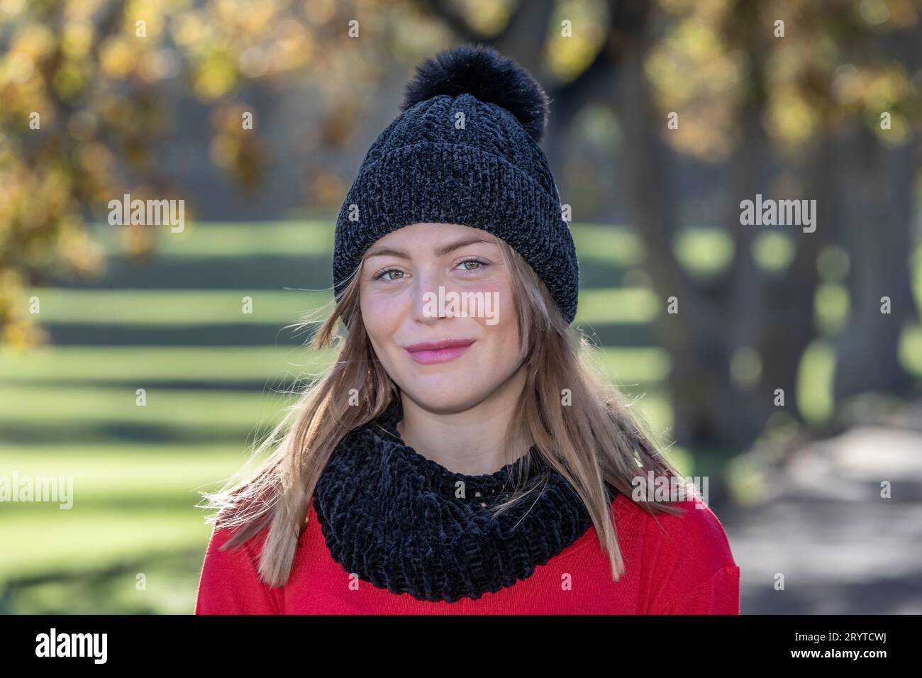 In un colpo alla testa e alle spalle, una splendida giovane donna bionda, che indossa un cappello di lana nera e un maglione rosso brillante, si irradia sotto gli alberi autunnali Foto Stock