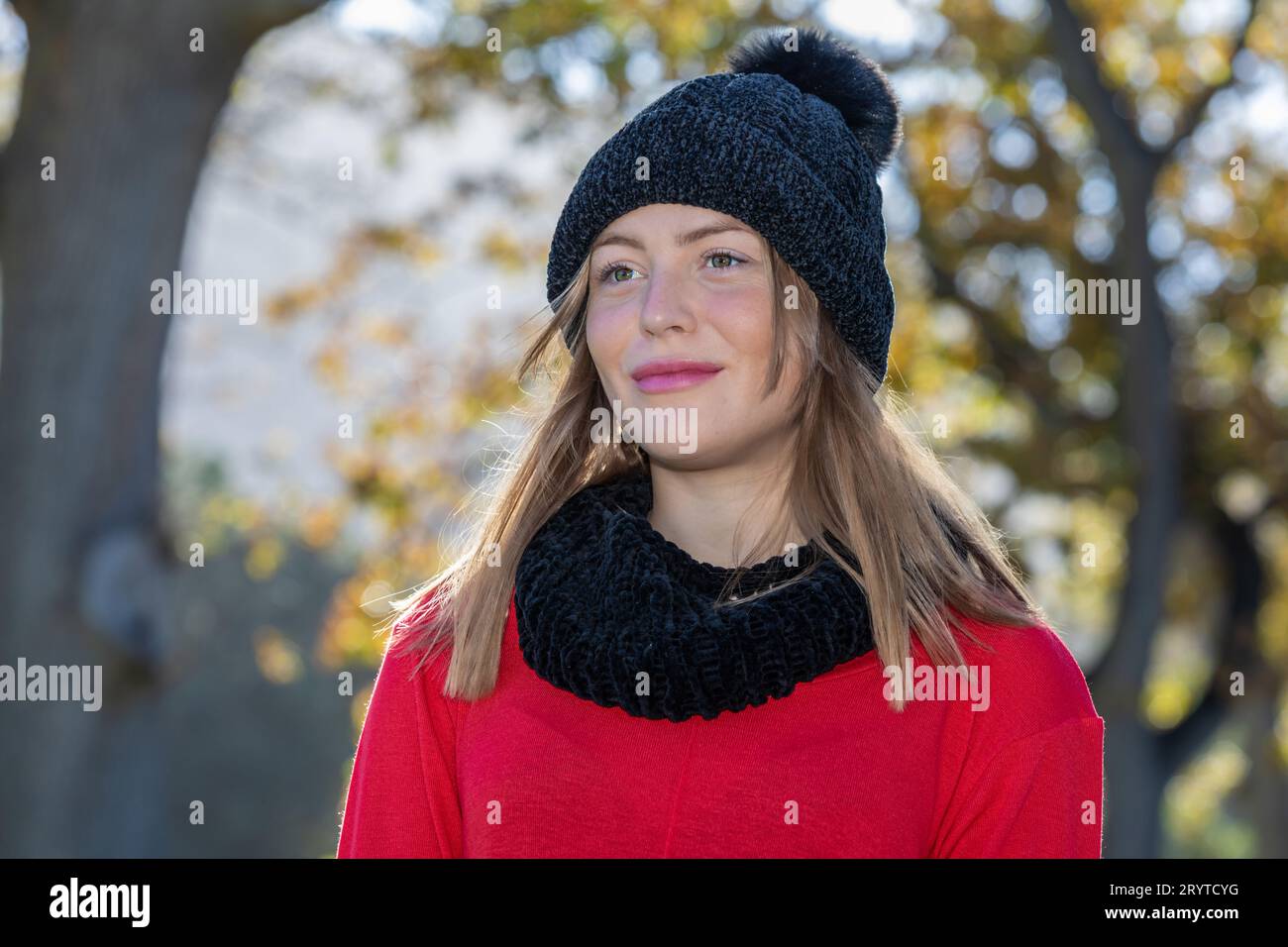 In un colpo alla testa e alle spalle, una splendida giovane donna bionda, che indossa un cappello di lana nera e un maglione rosso brillante, si irradia sotto gli alberi autunnali Foto Stock