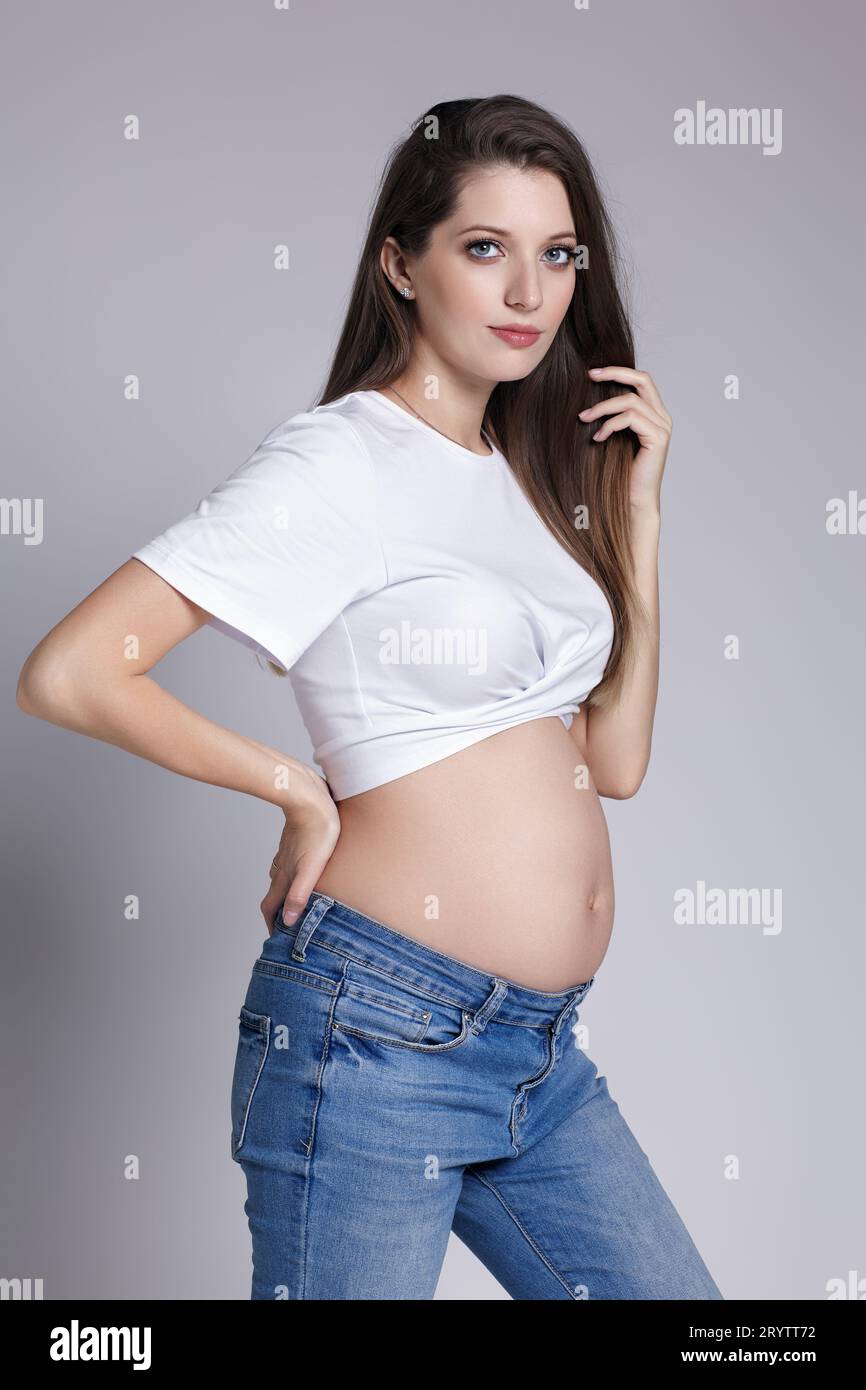 Giovane donna incinta in t-shirt bianca e jeans. su sfondo grigio. Pancia esposta. Donna su 5th mese di gravidanza. Foto Stock
