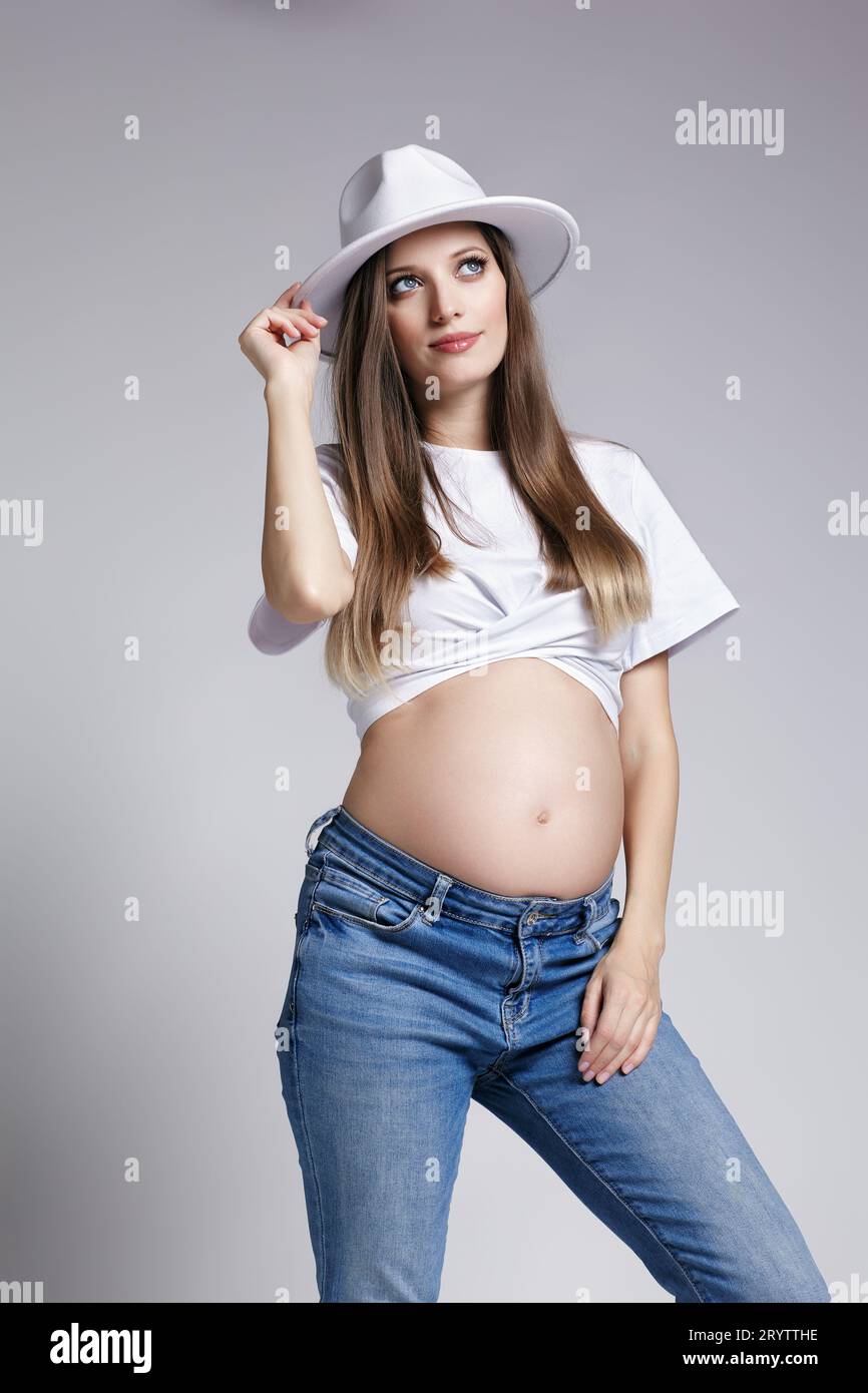 Giovane donna incinta con t-shirt bianca, cappello e jeans. Femmina con pancia esposta. quinto mese di gravidanza Foto Stock