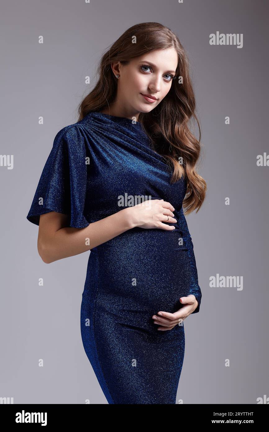 Ritratto di una giovane bella donna incinta su sfondo grigio studio. Donna in abito di paillettes blu con le mani vicino alla pancia incinta Foto Stock