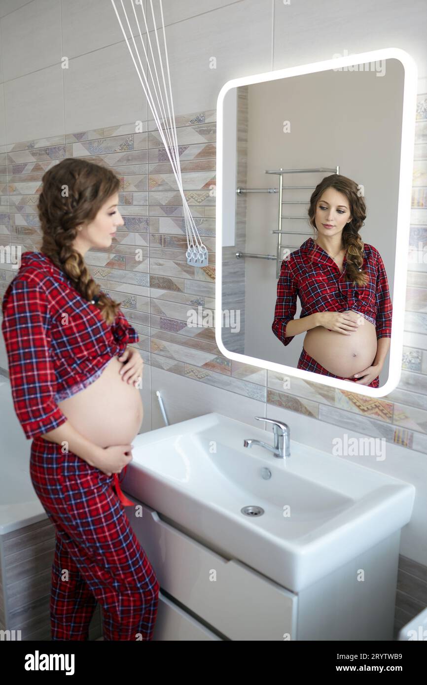 Una donna incinta nel bagno che guarda nello specchio. Donna vestita di pigiama rosso e pancia aperta che gode del suo stato di gravidanza Foto Stock