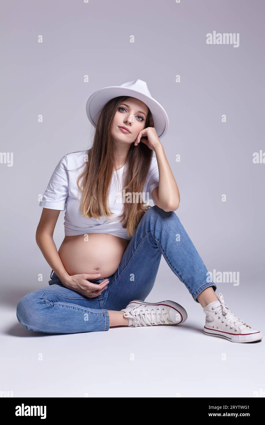 Giovane donna incinta con t-shirt bianca, cappello e jeans seduti sul pavimento sullo sfondo. Femmina con pancia esposta. quinto mese Foto Stock