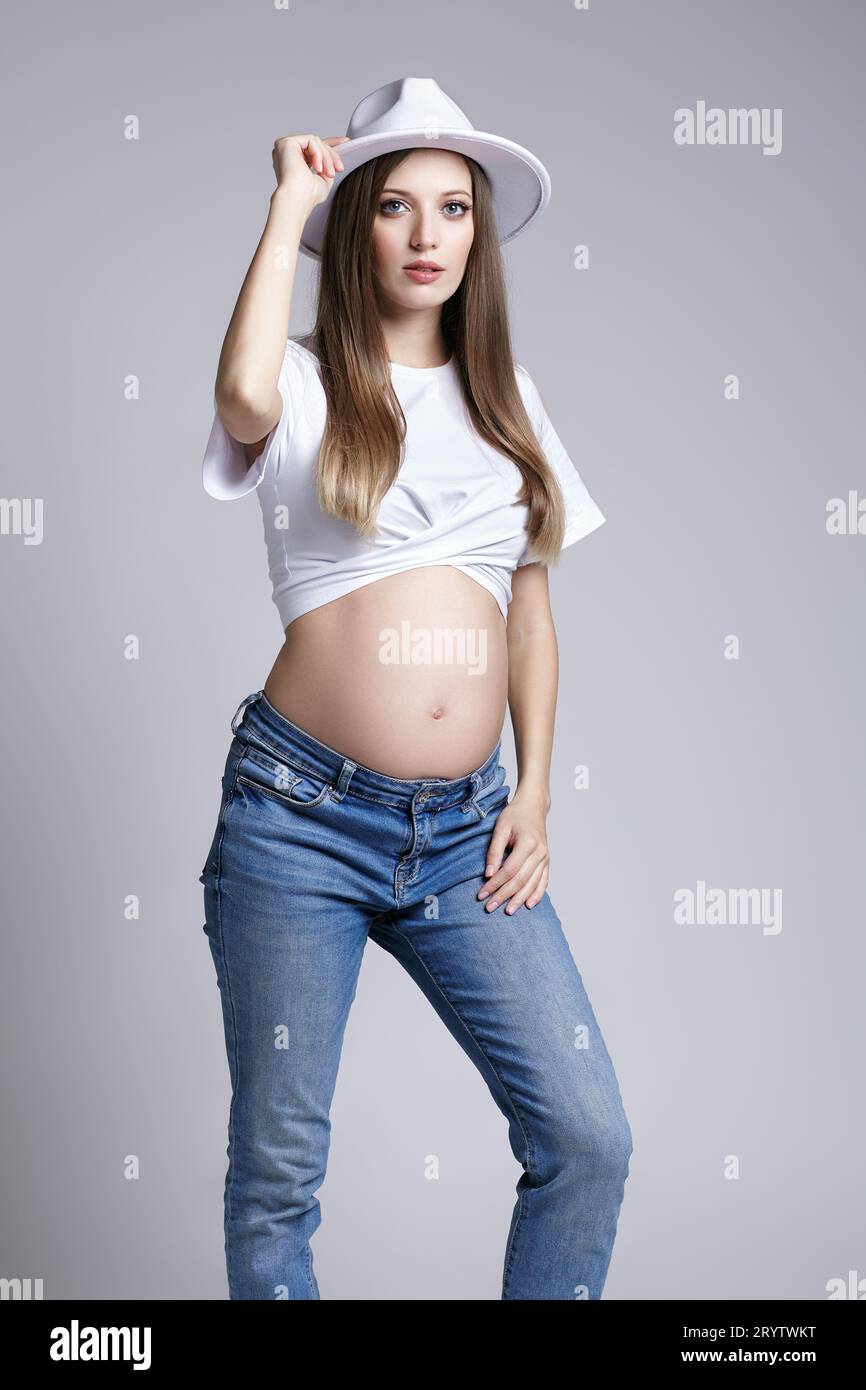 Giovane donna incinta con t-shirt bianca, cappello e jeans. Femmina con pancia esposta. quinto mese di gravidanza Foto Stock