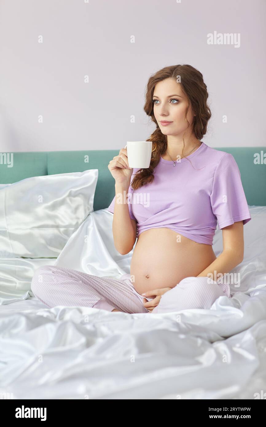 Una donna incinta giace nel letto della sua camera da letto domestica. Donna che gode del suo stato di gravidanza. Una tazza di tè in mano. Foto Stock
