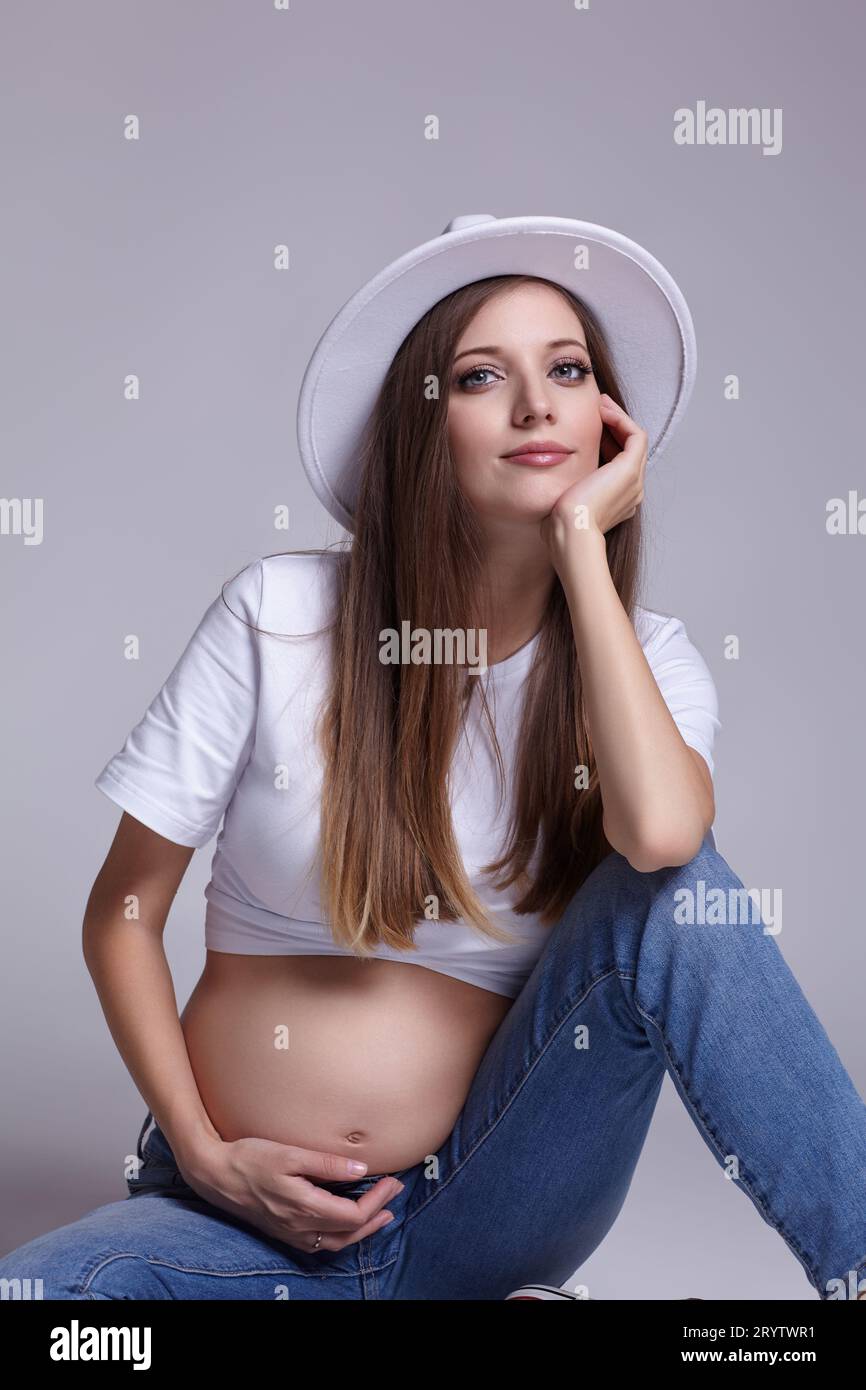 Giovane donna incinta con t-shirt bianca, cappello e jeans seduti sul pavimento sullo sfondo. Femmina con pancia esposta. quinto mese Foto Stock