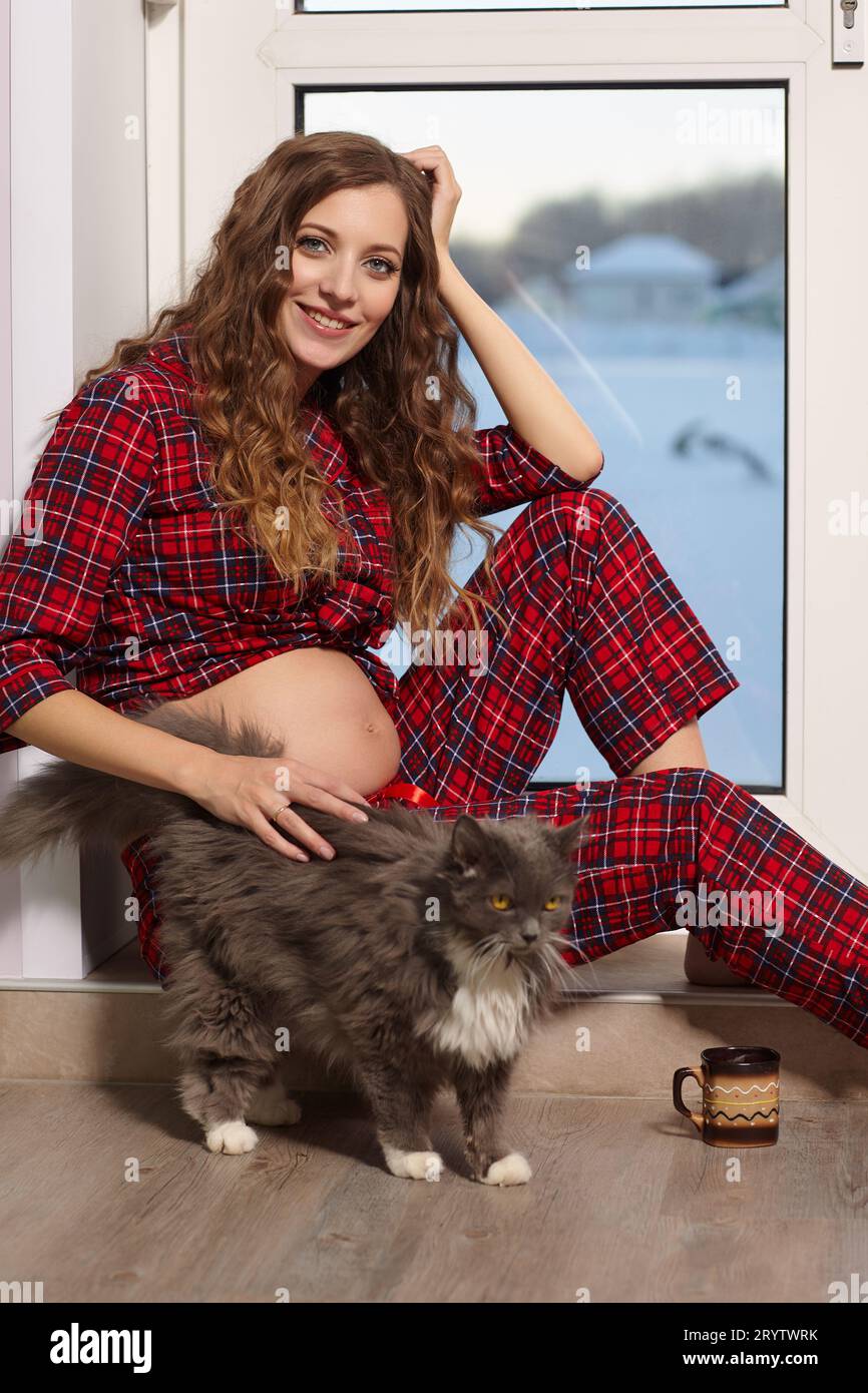 Una donna incinta nel soggiorno con una tazza di tè siede sulla soglia di una porta di vetro e gioca con il gatto. Foto Stock