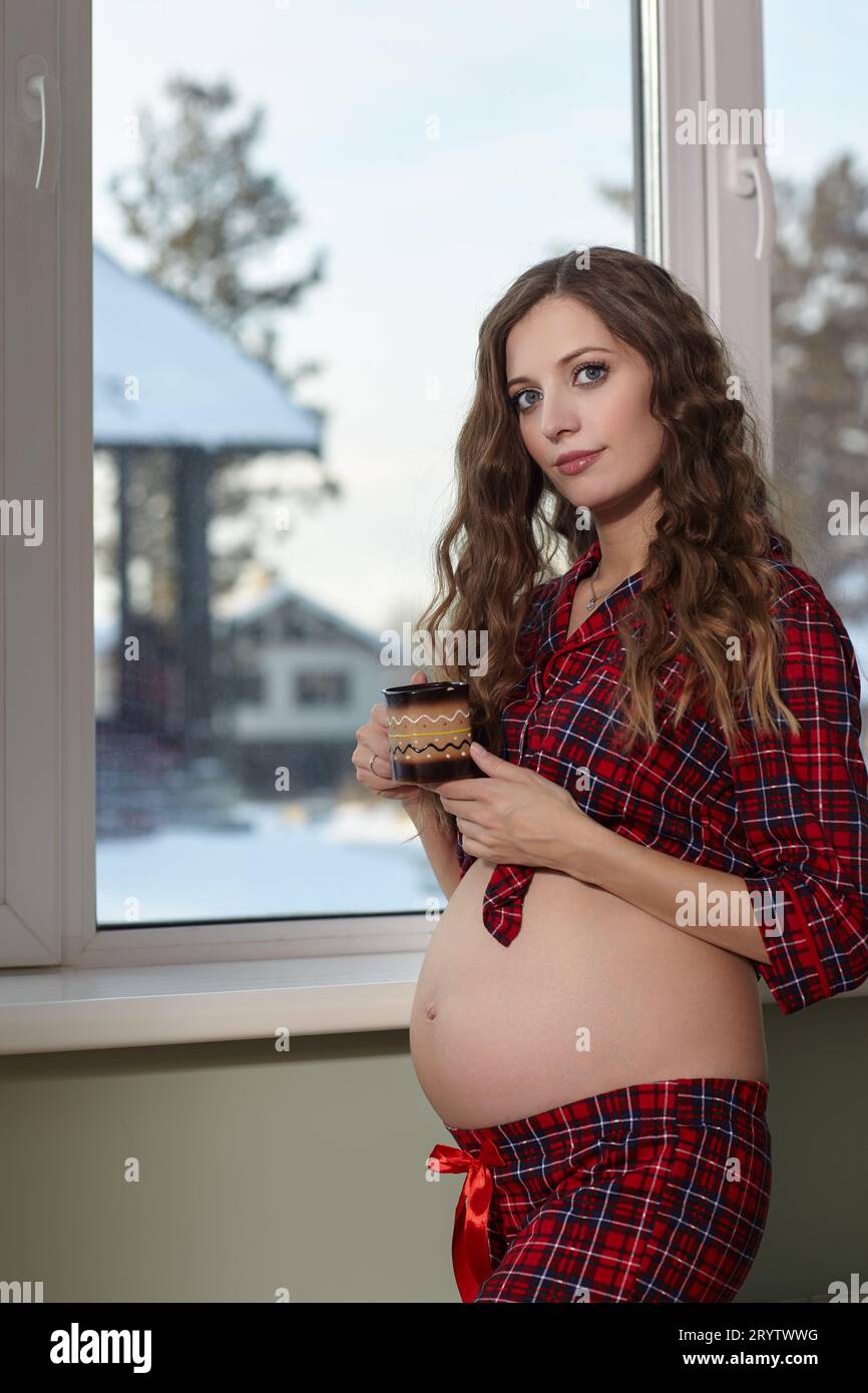 Una donna incinta nel soggiorno con una tazza di tè vicino alla finestra. Donna vestita di pigiami rosso e pancia aperta godendo il suo stato di gravidanza. Foto Stock