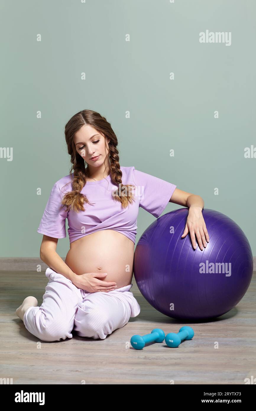 Una donna incinta siede vicino a una palla di fitness e guarda la sua pancia incinta. Foto Stock