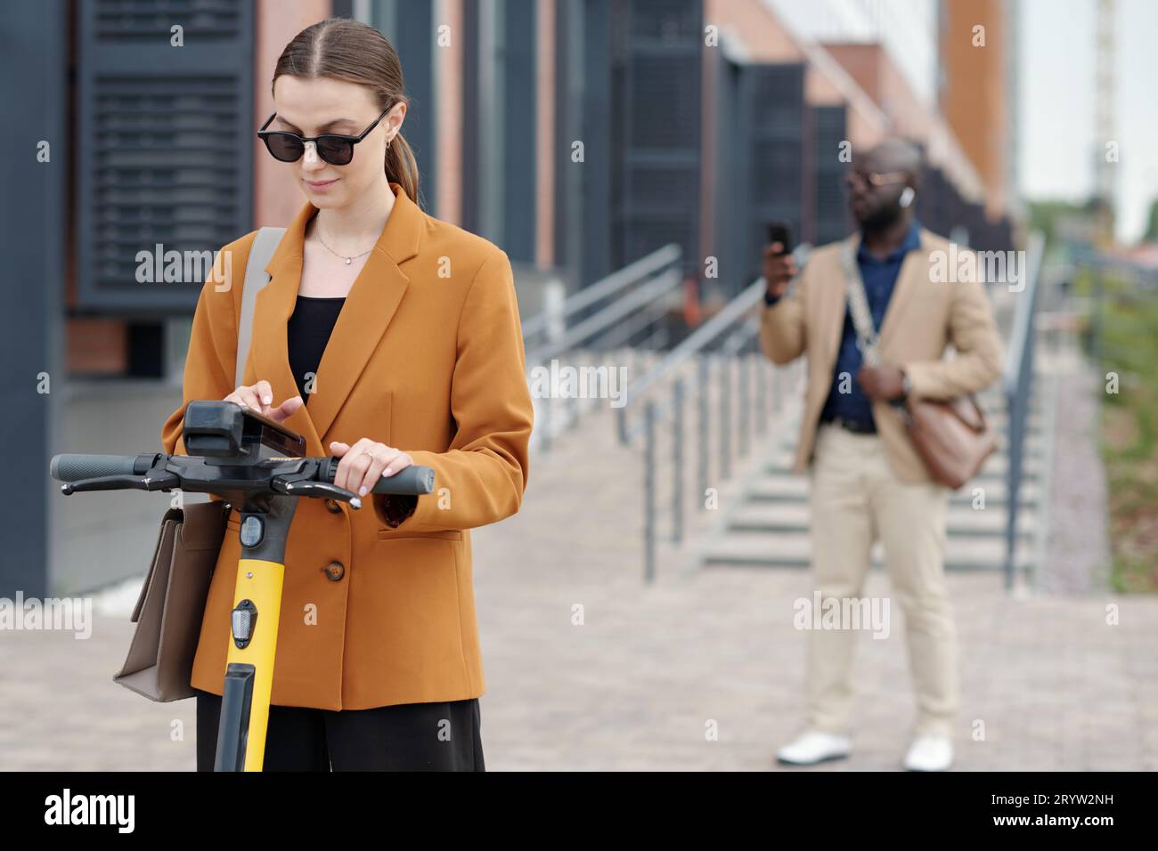 Giovane donna d'affari con occhiali da sole che sceglie le impostazioni di uno scooter elettrico mentre si trova di fronte alla macchina fotografica contro un collega afroamericano Foto Stock