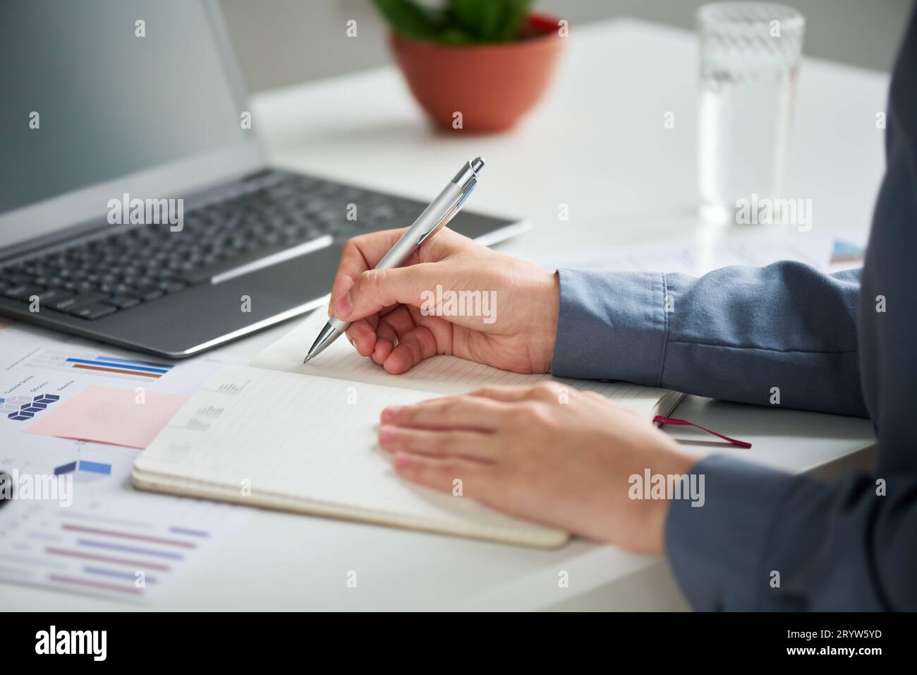 Concentratevi sulla mano di una giovane donna d'affari in camicia blu con penna su una pagina vuota del notebook durante l'organizzazione del lavoro e la stesura dell'agenda Foto Stock