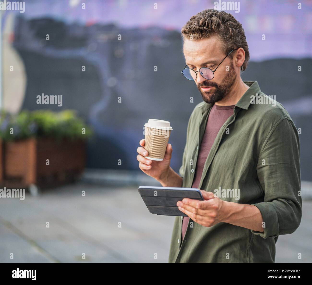 Uomo che si gode una pausa caffè all'aperto mentre legge informazioni dal suo tablet PC. L'atmosfera rilassata e serena si completa Foto Stock