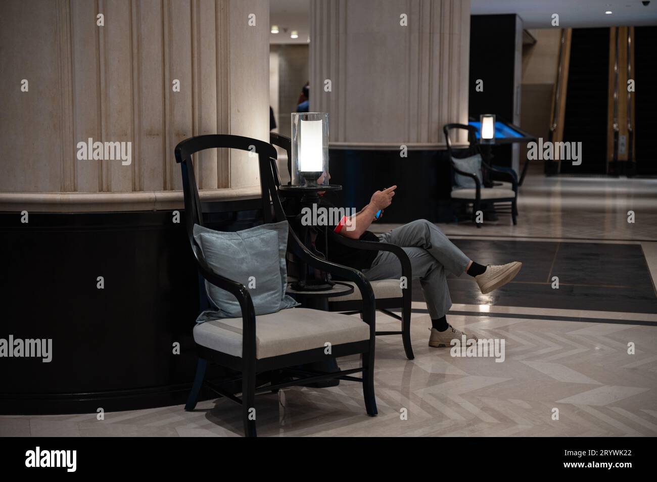 03.08.2023, Singapore, Repubblica di Singapore, Asia - Un ospite dell'hotel siede dietro una lampada nella lobby dello Shangri-la Hotel guardando il suo telefono. Foto Stock