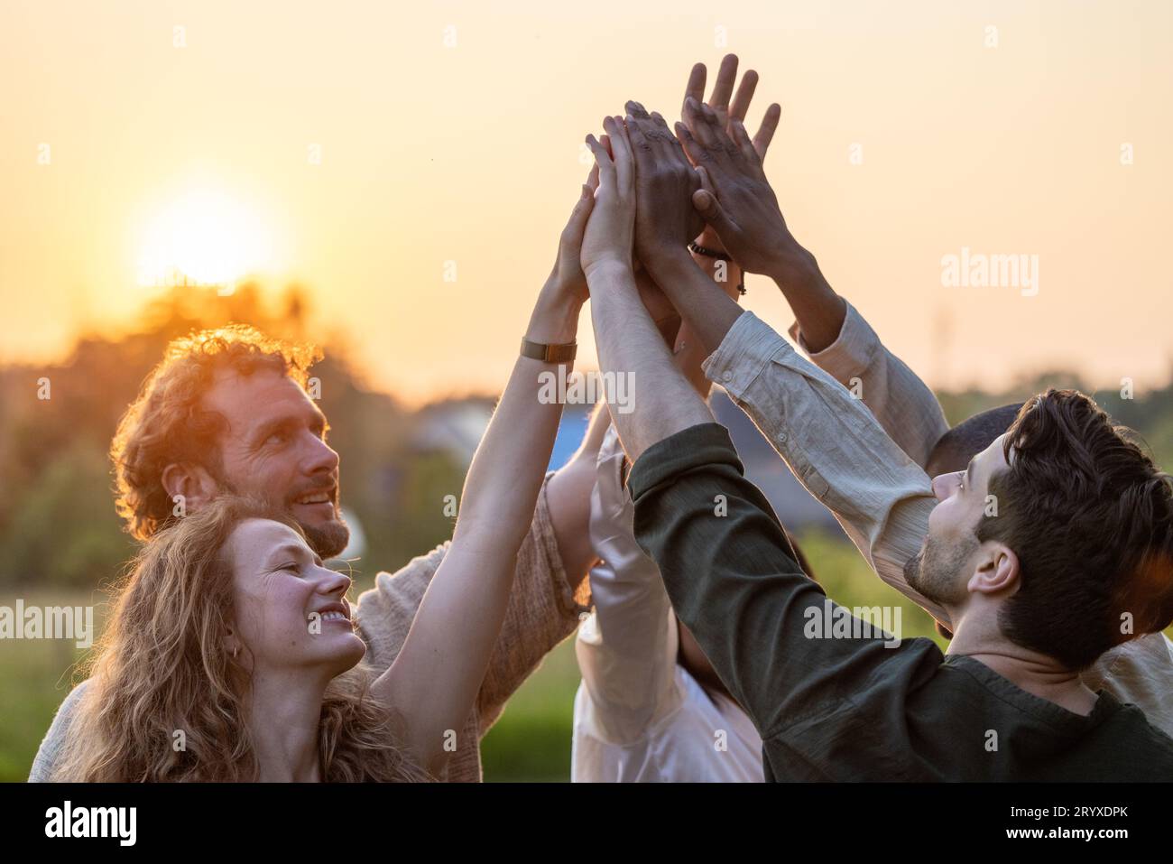 Gruppo multirazziale di giovani che danno cinque in alto contro un sole che tramonta, si sente entusiasta di concentrarsi da vicino sulle palme impilate Foto Stock