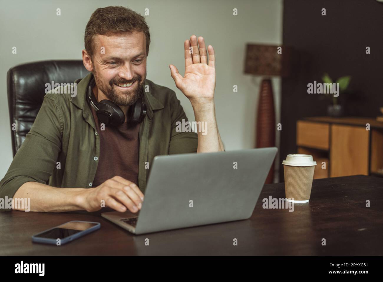 Uomo professionista impegnato in riunioni online in un ufficio loft. Con un gesto amichevole, saluta virtualmente qualcuno, dimostrati Foto Stock