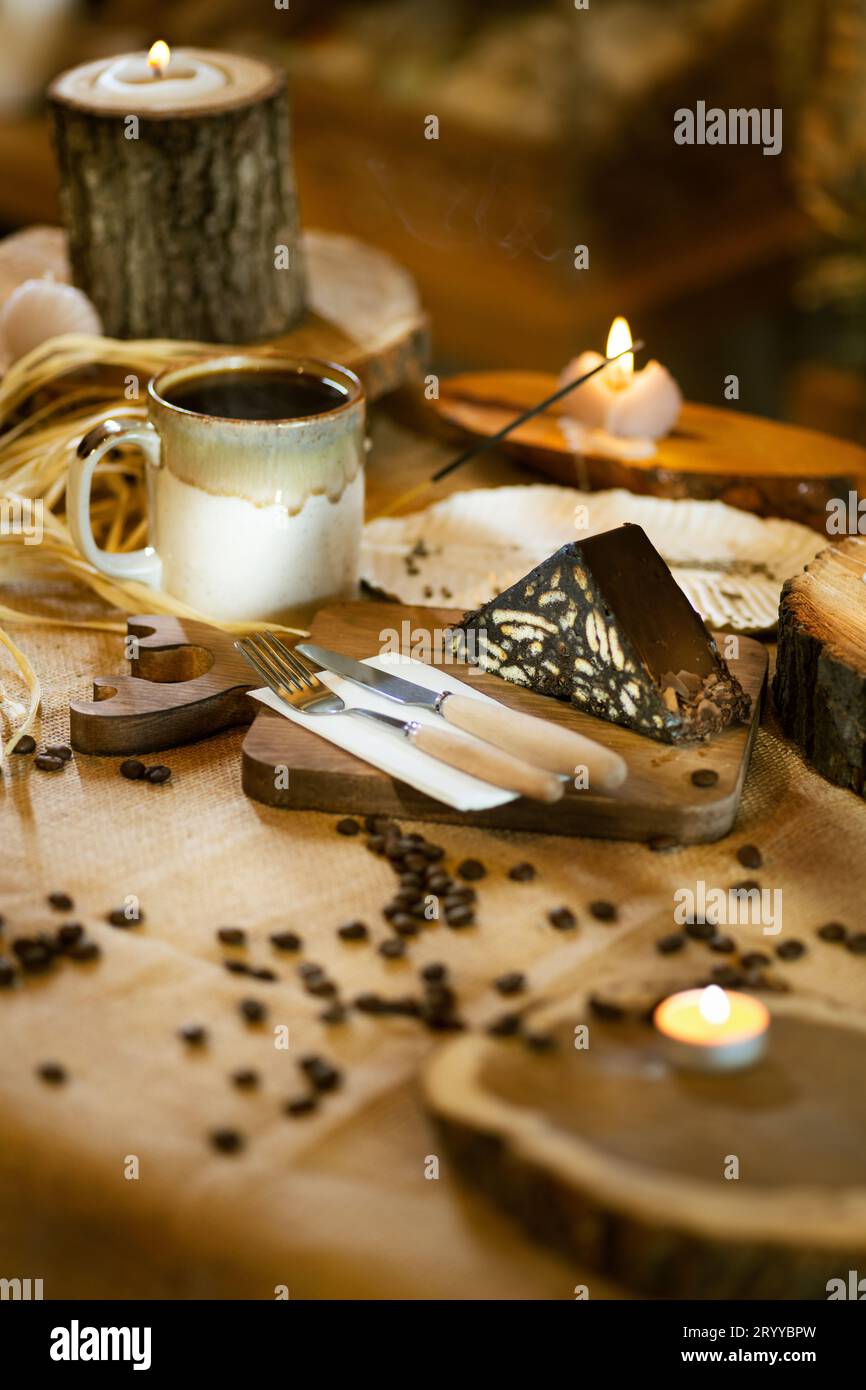 Torta a mosaico e caffè serviti su burlap, con posate e tavola di legno. Arricchito da incenso e atmosfera a lume di candela. Foto Stock