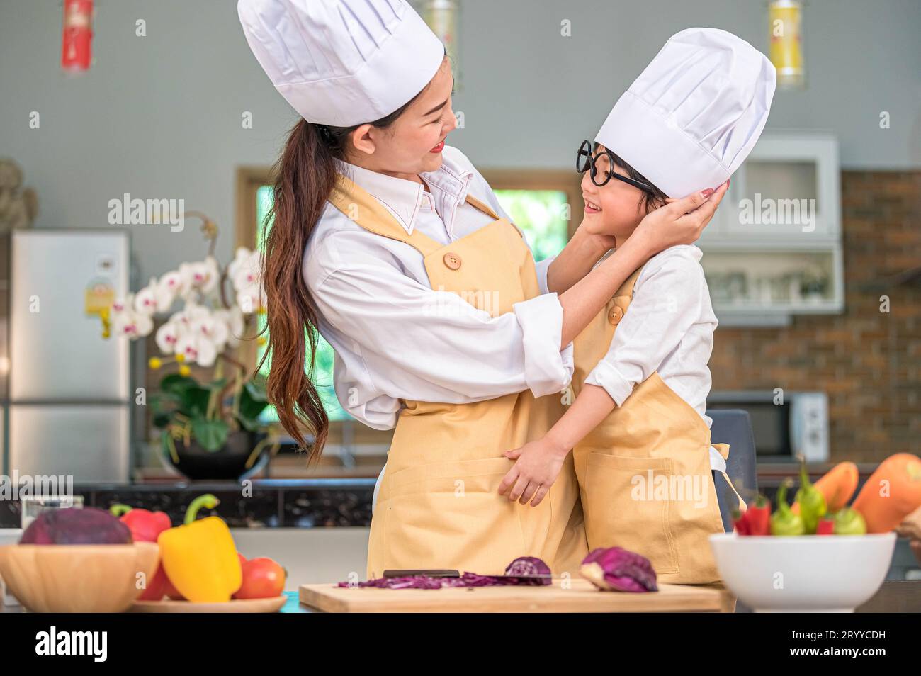 Un bambino felice e carino con occhiali da vista che sembrano bellissime donne asiatiche, madri a vicenda, mentre si preparano a cucinare in cucina all'hom Foto Stock