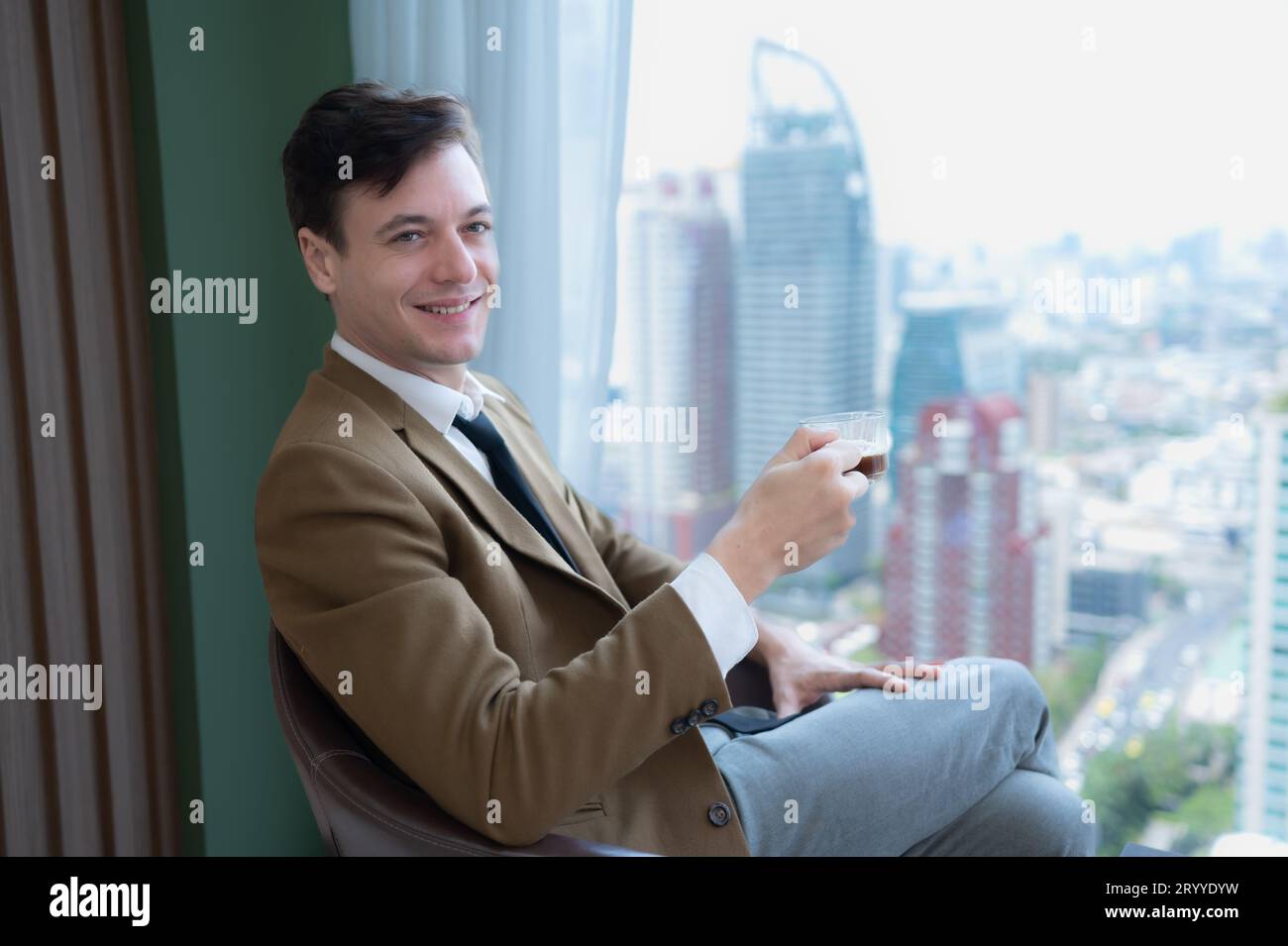 Giovane uomo d'affari seduto e rilassarsi nella sala relax vicino alla finestra che si affaccia sugli splendidi edifici della città. Foto Stock