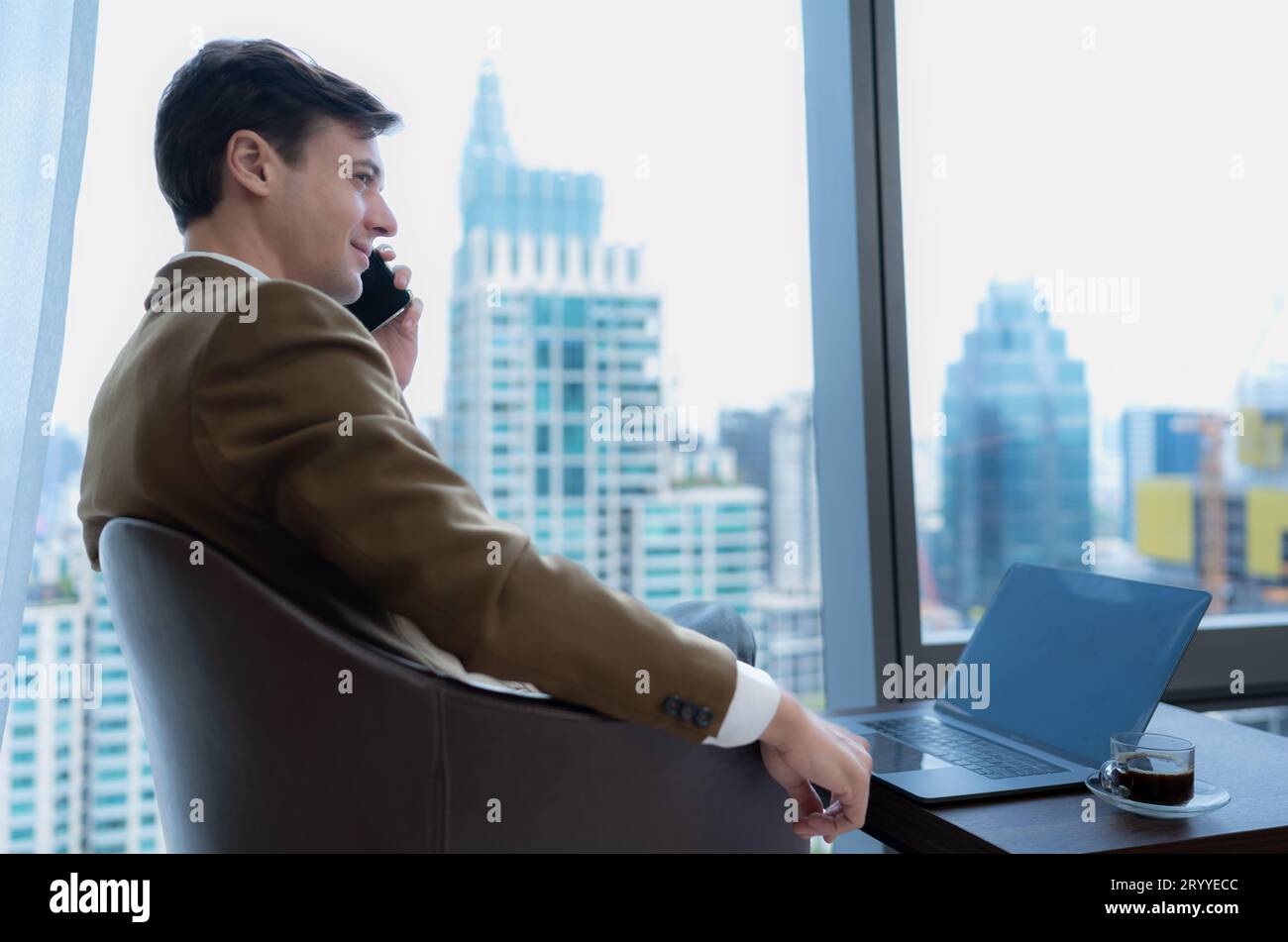 Giovane uomo d'affari seduto e rilassarsi nella sala relax vicino alla finestra che si affaccia sugli splendidi edifici della città. insieme al ph Foto Stock