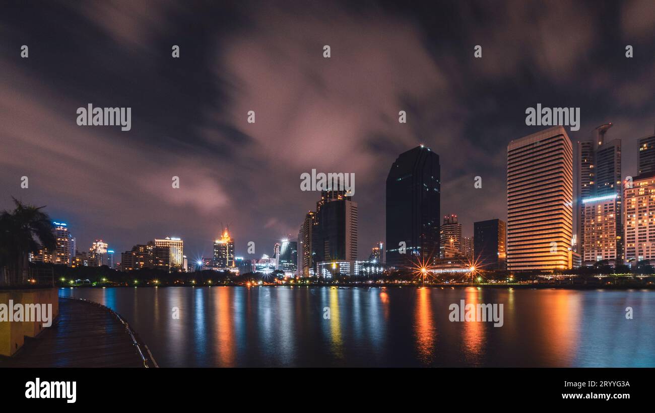 Grande città nella vita notturna con riflessi di onde d'acqua. Tecniche di esposizione lunga. Panorama del paesaggio. Concetto urbano e cittadino. Foto Stock