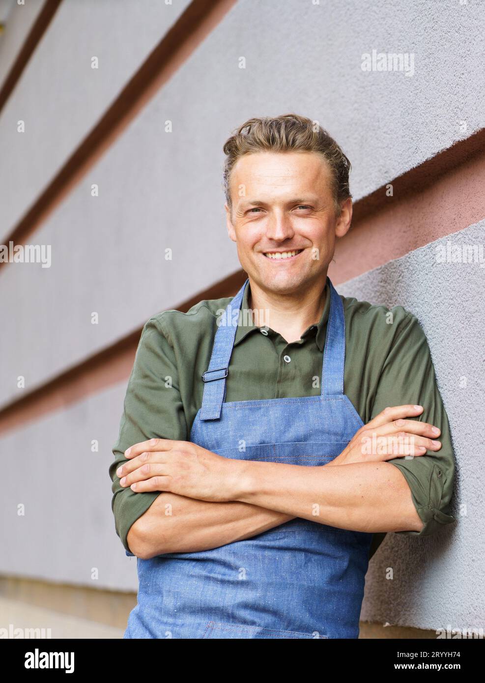 Cameriere esperto e dedicato o addetto al servizio in ambiente ristorante. Vestito in grembiule e uniforme, si trova accanto al muro Foto Stock