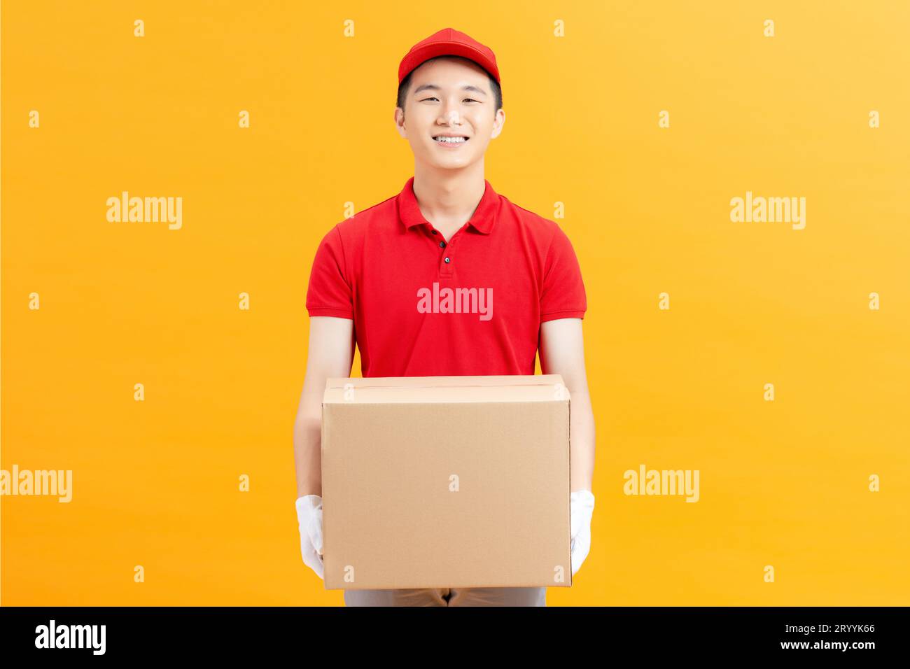 Un giovane e felice addetto alla consegna con tappo rosso in piedi con una cassetta postale isolata su sfondo giallo. Foto Stock