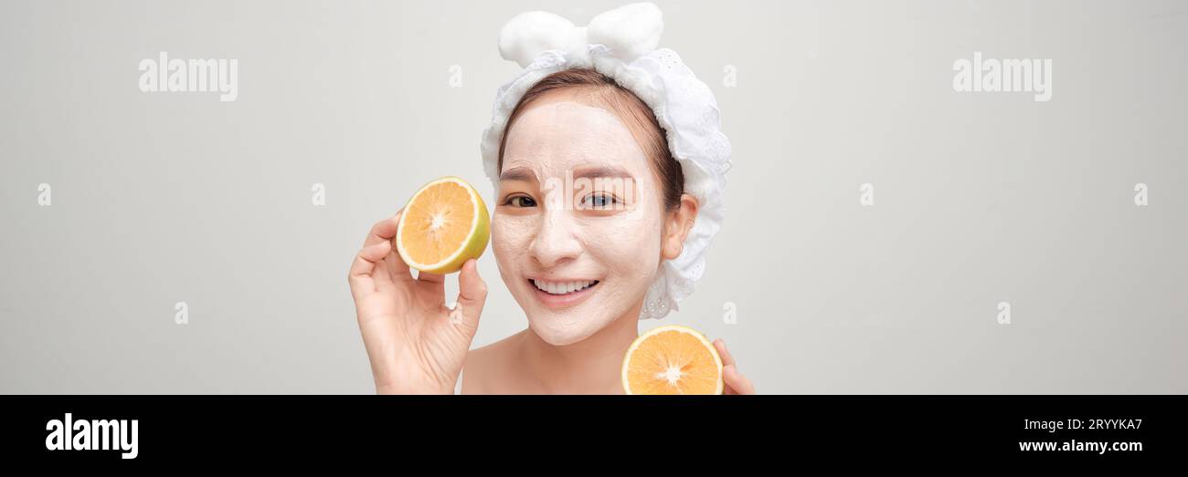Giovane donna con maschera facciale in argilla che tiene fetta di frutta arancione che copre gli occhi su striscione bianco Foto Stock