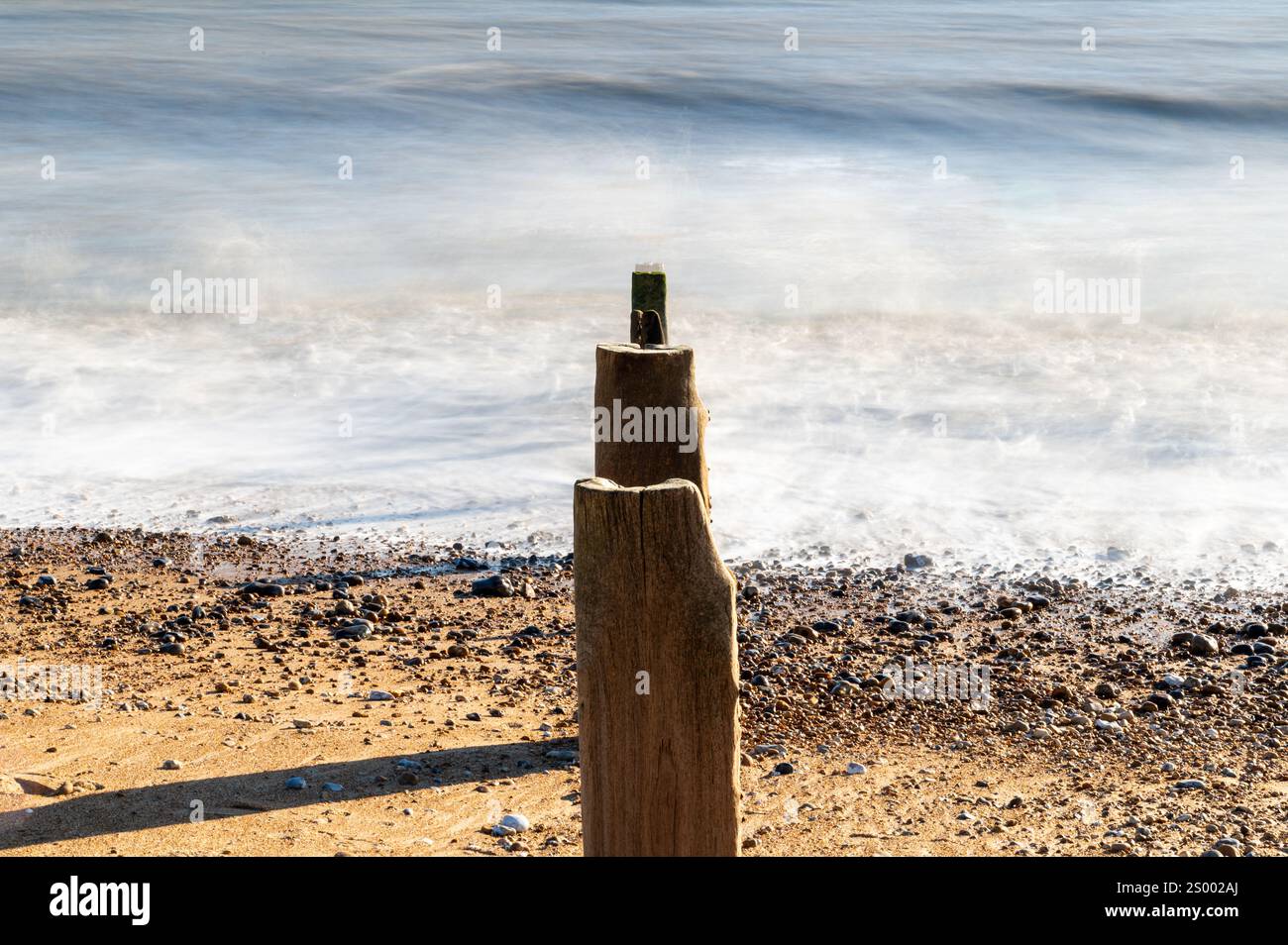 Foto a lunga esposizione di pali di groyne in fila a Bexhill-on-Sea, East Sussex, con onde lisce e fumose contro la costa in una soleggiata giornata invernale Foto Stock